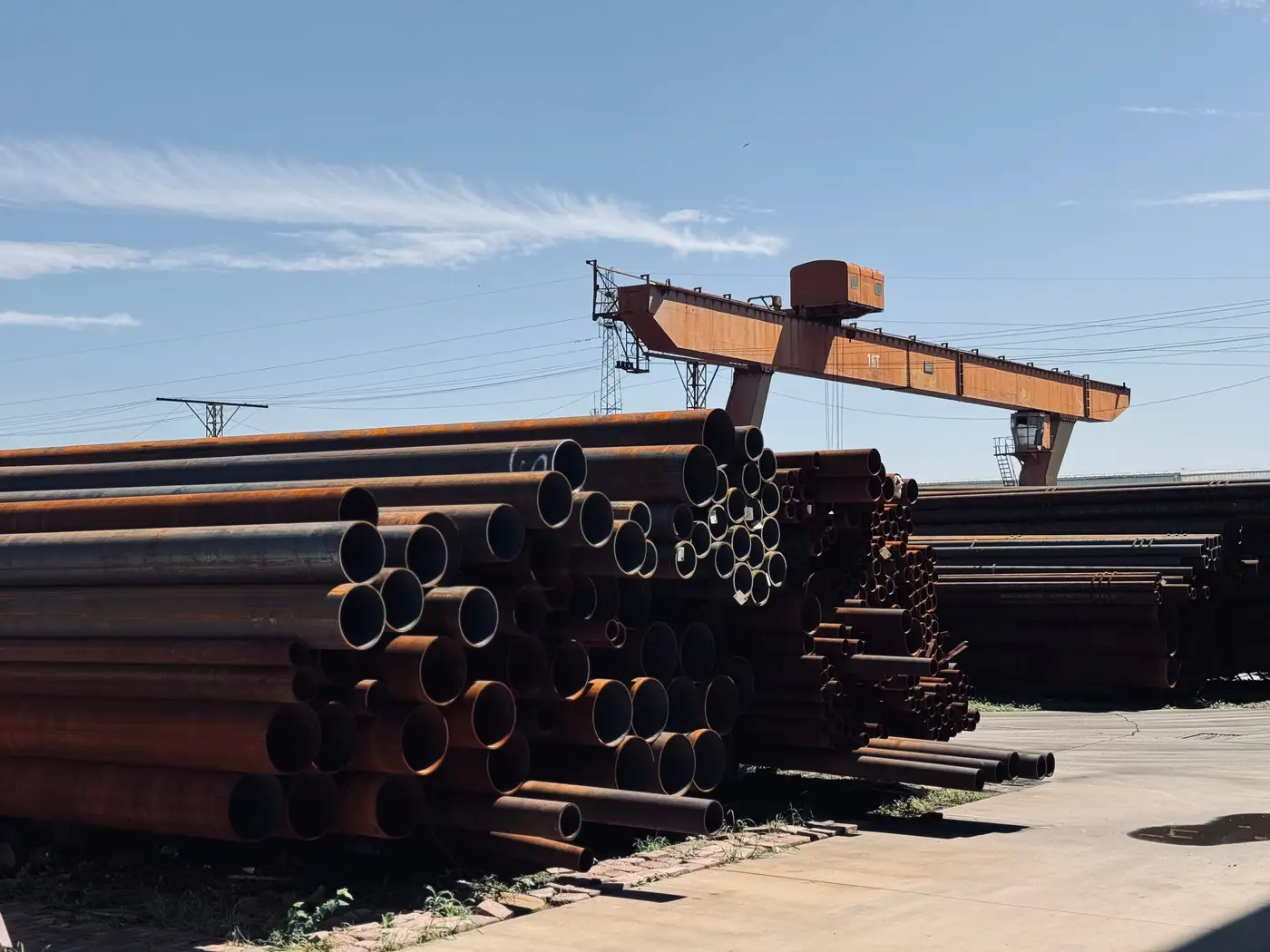Stacked steel pipes and an overhead gantry crane in an outdoor industrial storage yard.