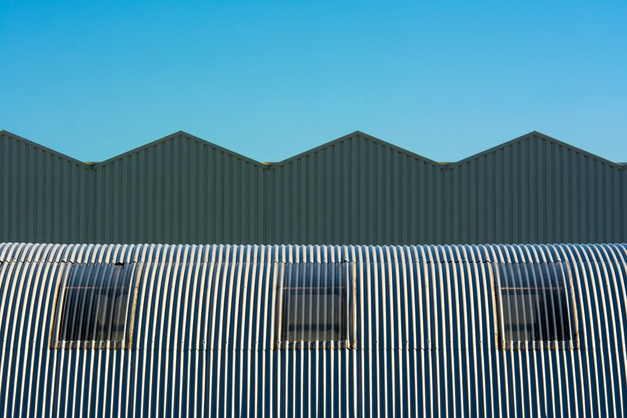 Sawtooth factory roof rising behind a curved corrugated shed roof under a clear sky